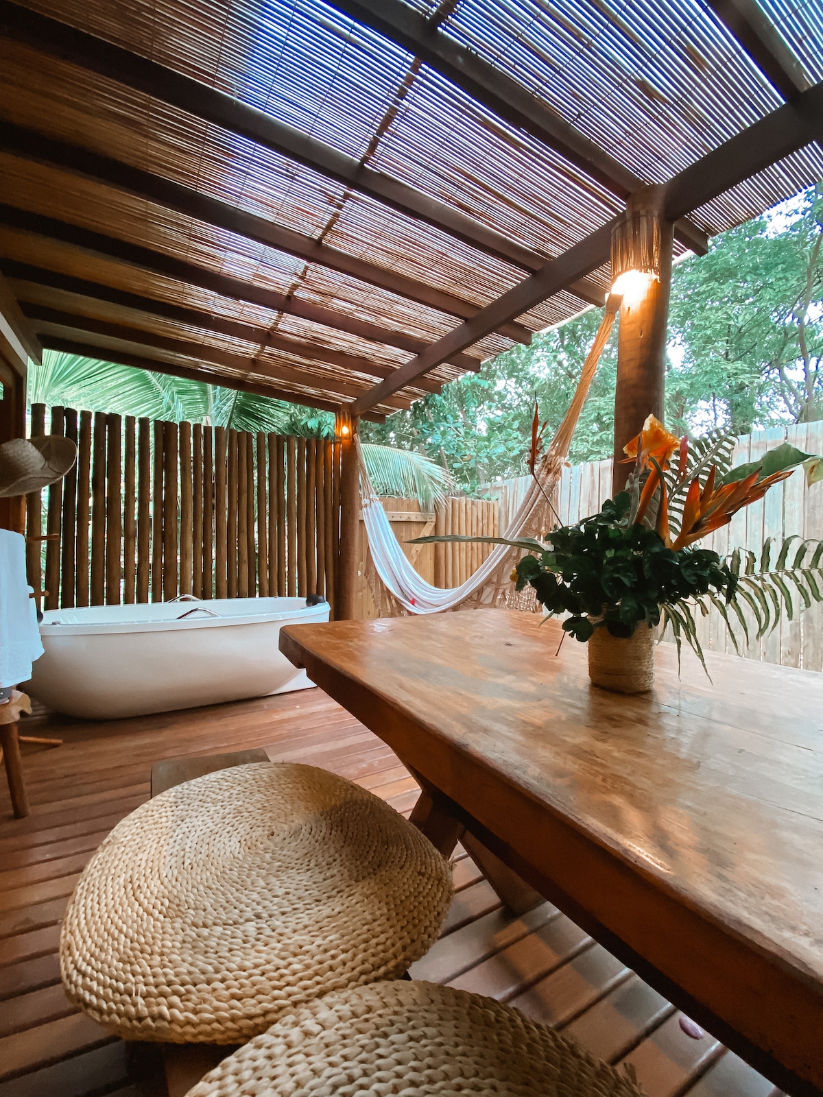 An outdoor seating area features a wooden table accompanied by two round, natural fiber placemats. A hammock is draped nearby, and a freestanding bathtub is positioned under a partially covered roof. Green foliage is visible in the background, enhancing the peaceful ambiance.