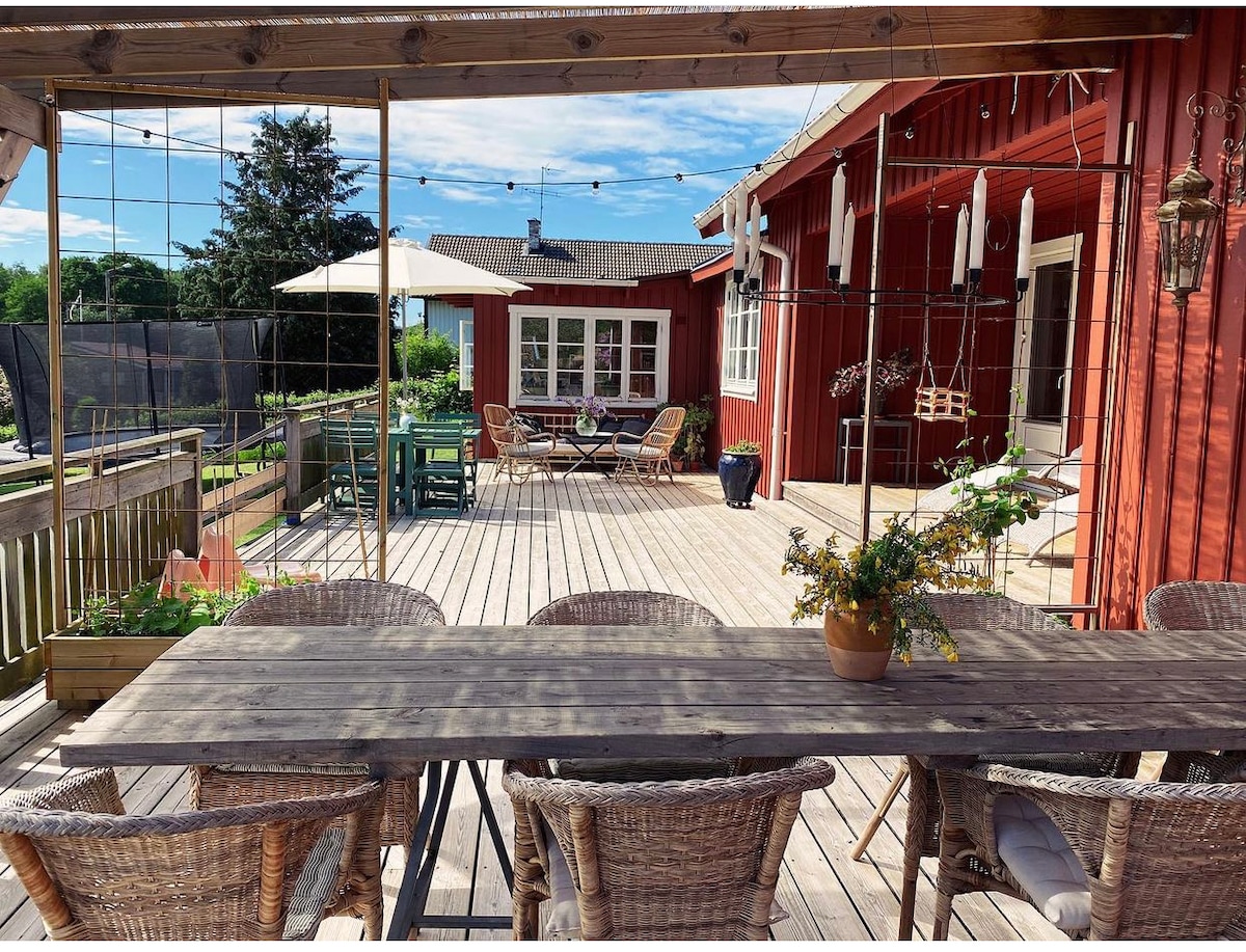 A spacious outdoor deck features a long wooden dining table surrounded by woven chairs. Various seating areas are present, including a set of comfortable lounge chairs and a parasol for shade. String lights hang above, creating a welcoming ambiance.