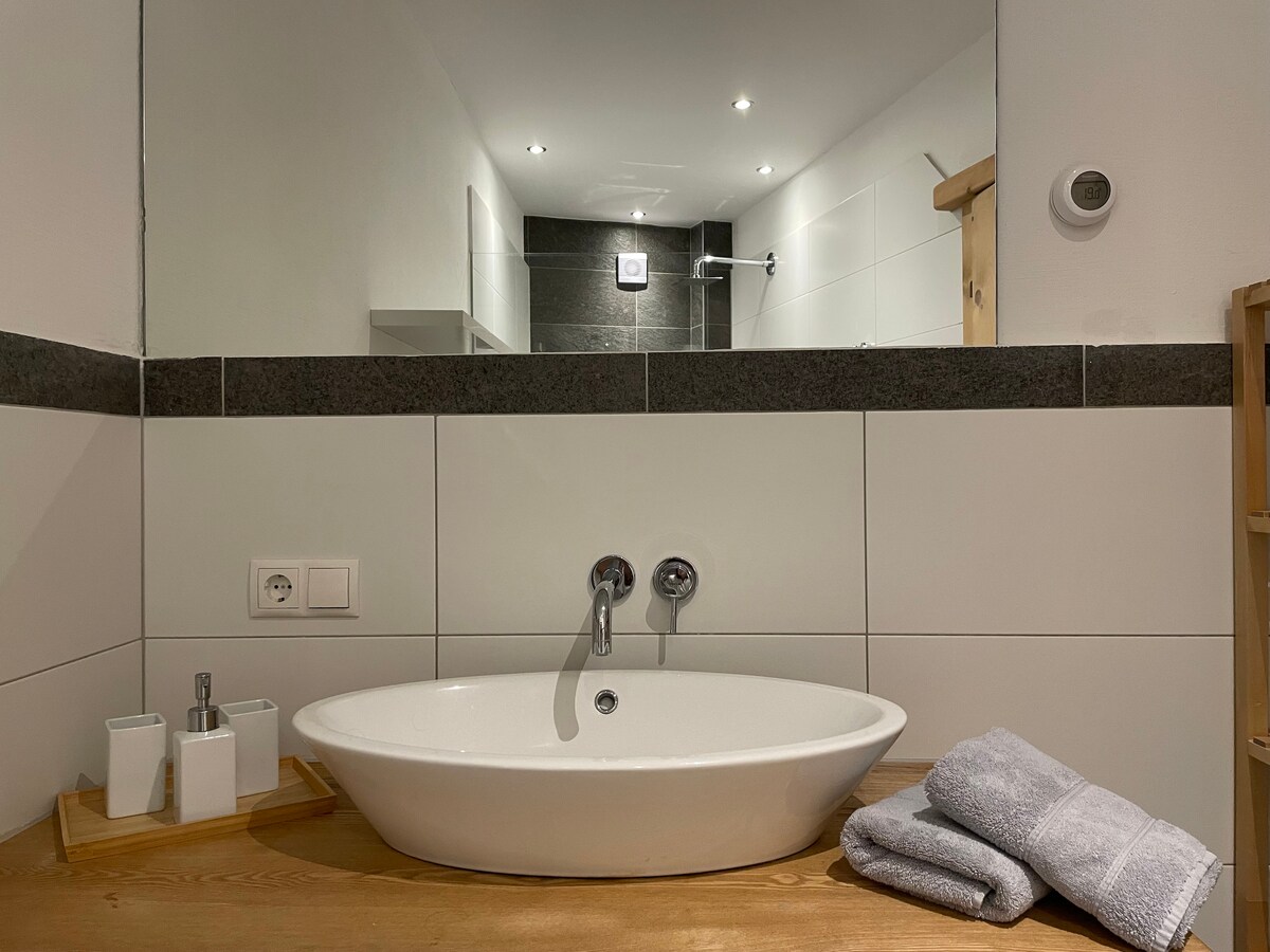 A modern bathroom is featured, showcasing a freestanding oval sink set on a wooden surface. Two neatly folded towels are placed nearby, and simple toiletries are arranged in containers. Soft lighting enhances the clean, minimalist design, with a large mirror reflecting the space.