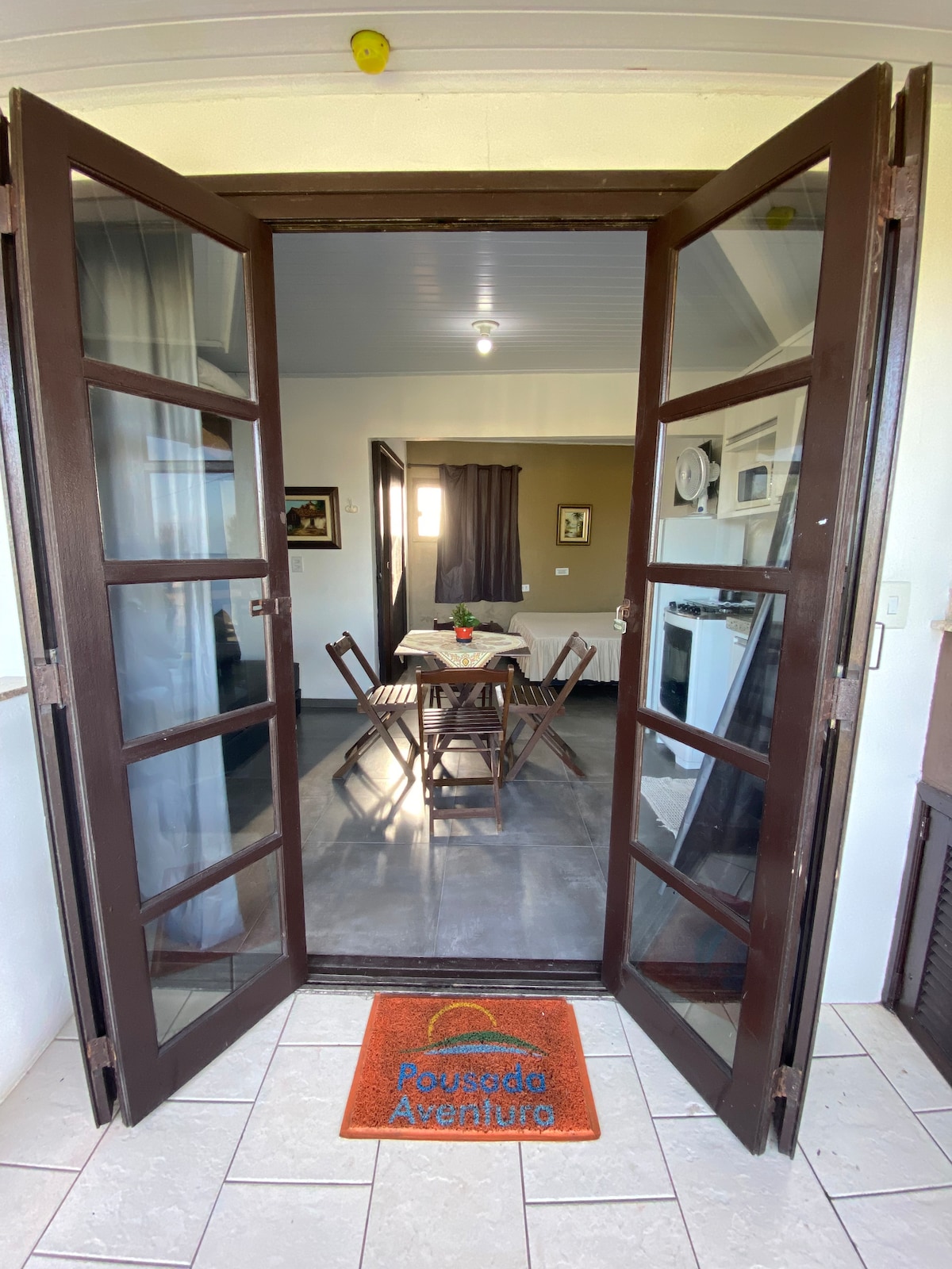 Double doors welcome guests into a bright dining area furnished with a table and wooden chairs. A small decorative centerpiece sits on the table, while kitchen appliances are visible in the background. Natural light enters through large windows, enhancing a fresh and open atmosphere.