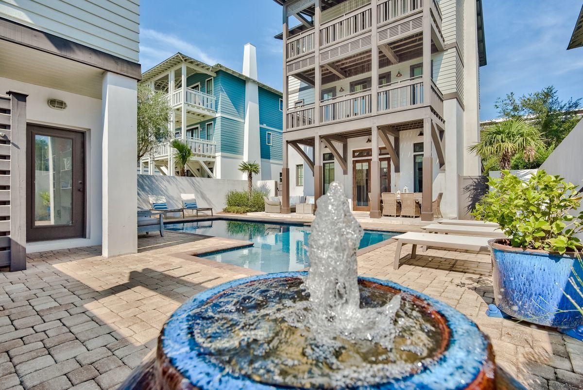 A tranquil courtyard showcases a private heated pool surrounded by natural stone. A sparkling fountain is positioned in the foreground, with poolside lounge chairs and a spacious outdoor seating area visible in the background, framed by a three-story home.