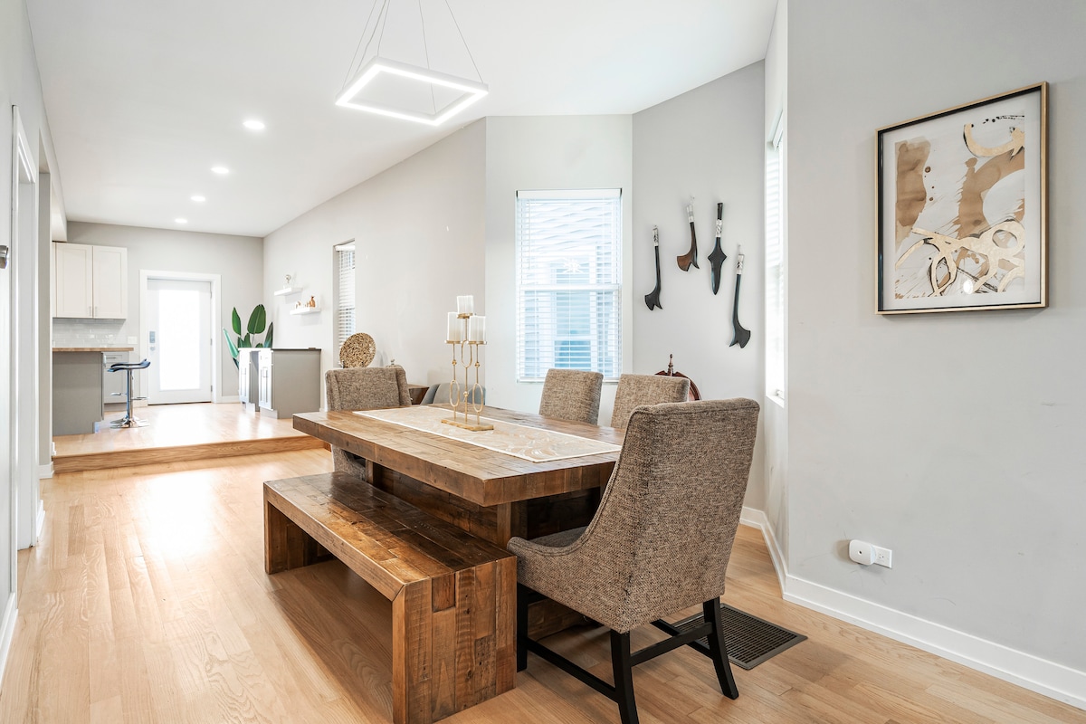 A dining area features a large wooden table accompanied by four upholstered chairs. Natural light filters through the windows, illuminating the space. Decor accents include wall art and a small plant, enhancing the inviting feel of the room.