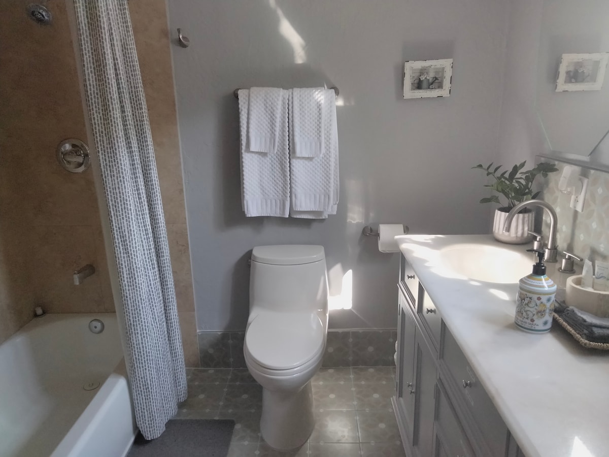 The bathroom features a modern design with light grey walls and natural stone accents. A white toilet is positioned next to a marble-topped vanity, which includes a sink and a decorative soap dispenser. Soft towels are neatly hung on a rack, while natural light brightens the space.