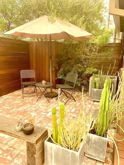 An outdoor sitting area is framed by wooden fencing, featuring two chairs and a small table under a beige umbrella. Planters with cacti are placed around the space, providing greenery against a brick patio.
