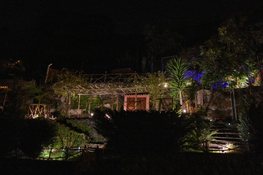 The property is set against a dark backdrop, illuminated by garden lights that highlight the greenery and stone structure. Various plants and trees surround the area, while a wooden terrace adds charm to the outdoor space.