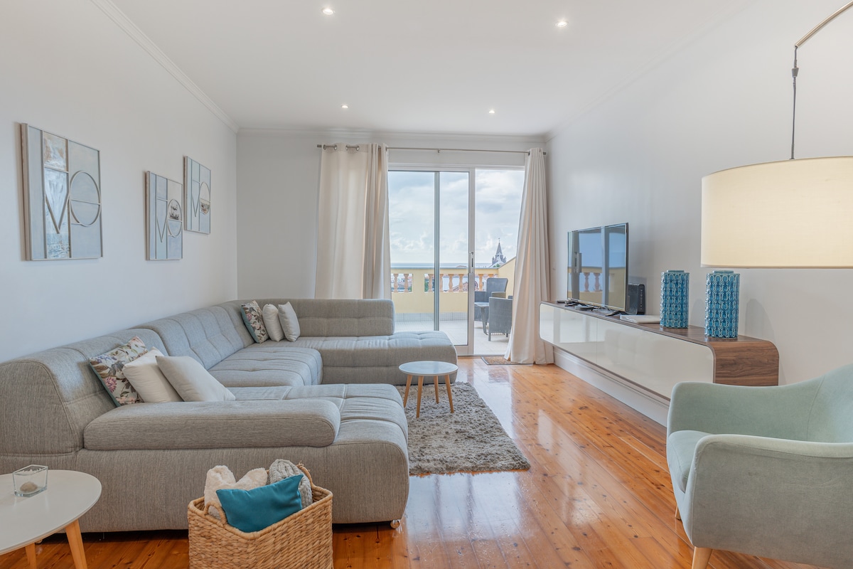 A spacious living area features a light grey sectional sofa complemented by decorative cushions. Natural light enters through large sliding doors leading to the outdoor space. A light wooden coffee table rests on a soft area rug, alongside a woven basket and a green accent chair.