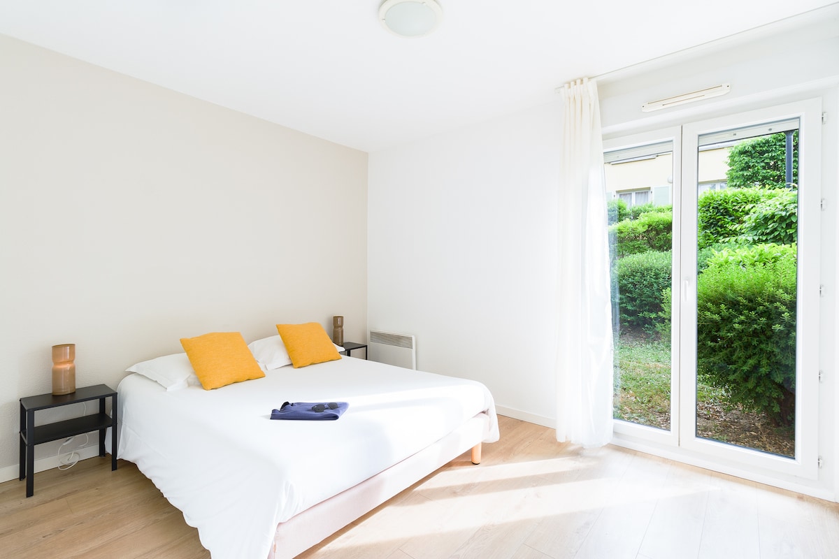 A spacious bedroom features a large bed dressed with a white duvet and two yellow accent pillows. A bedside table is situated on each side of the bed. Natural light fills the room through a pair of glass doors opening to a green outdoor space.