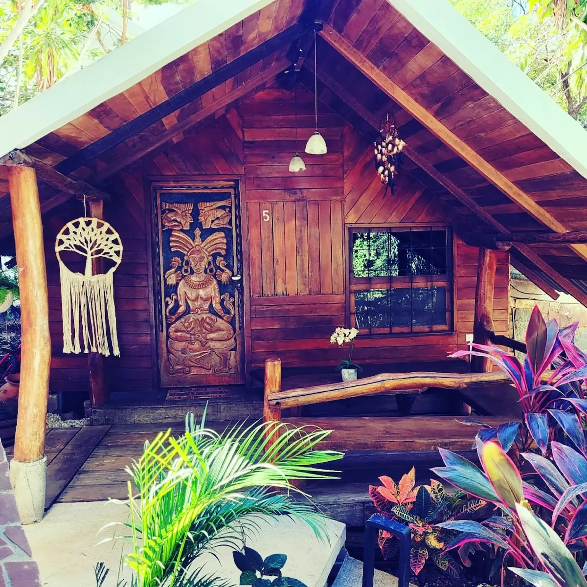 A quaint wooden cabin features a richly textured facade and a decorative wooden door with intricate carvings. Outdoor seating is provided on a bench made from natural wood, surrounded by lush green plants and vibrant flowers, creating a serene entrance atmosphere.