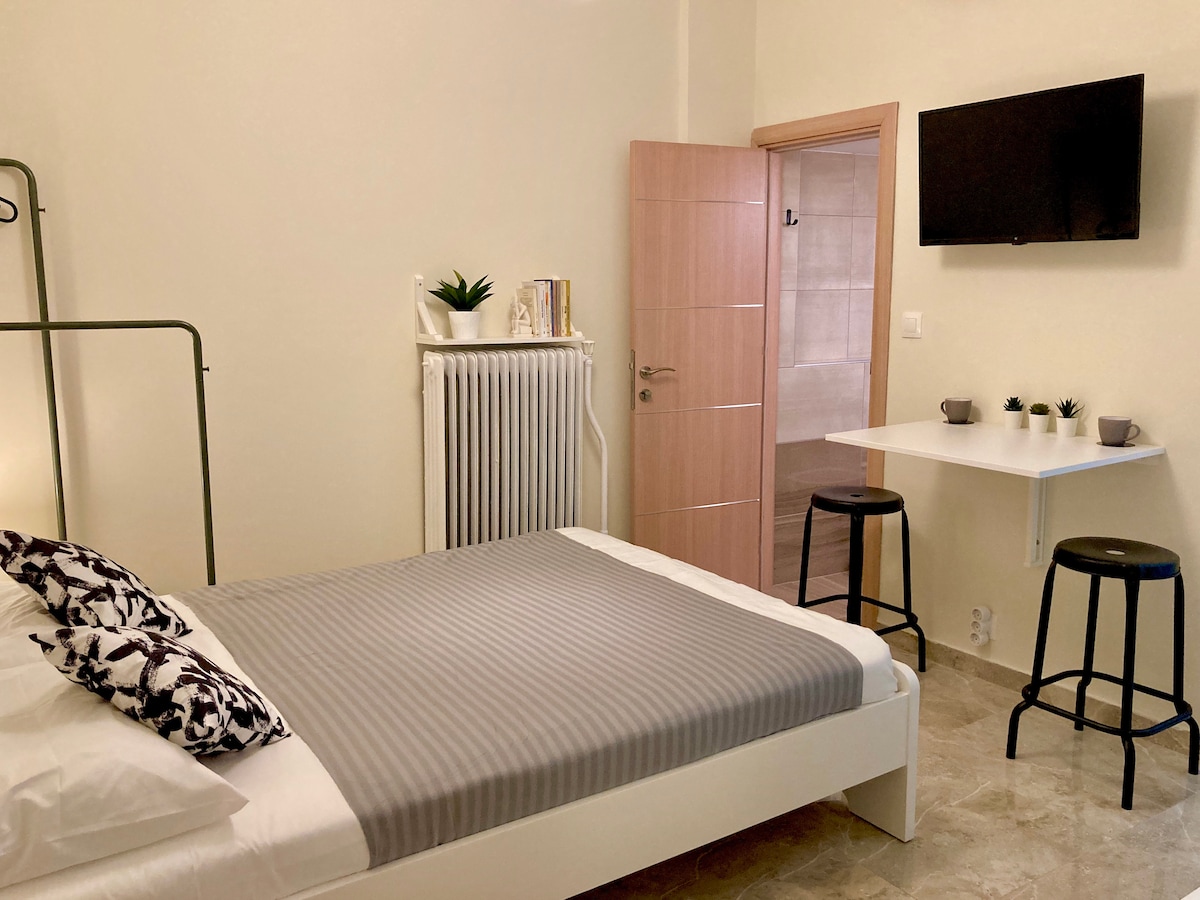 A neatly arranged sleeping area features a spacious bed with crisp white linens and a gray striped blanket. A small table with two black stools is positioned nearby, and a wall-mounted television is visible above. A door leads to a separate room with light-colored walls.