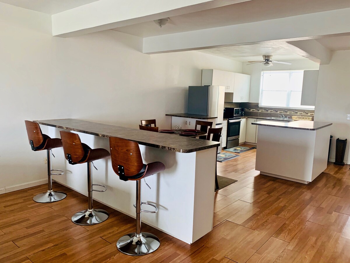 The spacious kitchen area is shown with modern appliances, including a refrigerator and stove. A large kitchen island with four bar stools enhances the social atmosphere. Light wood flooring complements the neutral color scheme, and natural light enters through the windows.