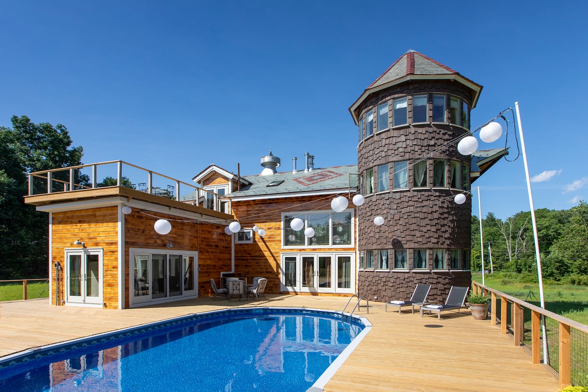 A large outdoor space features a swimming pool with lounge chairs nearby. The two-story structure showcases a unique tower and a wooden exterior, complemented by large windows and symmetrical lighting fixtures. A spacious deck surrounds the pool, offering views of the surrounding greenery.