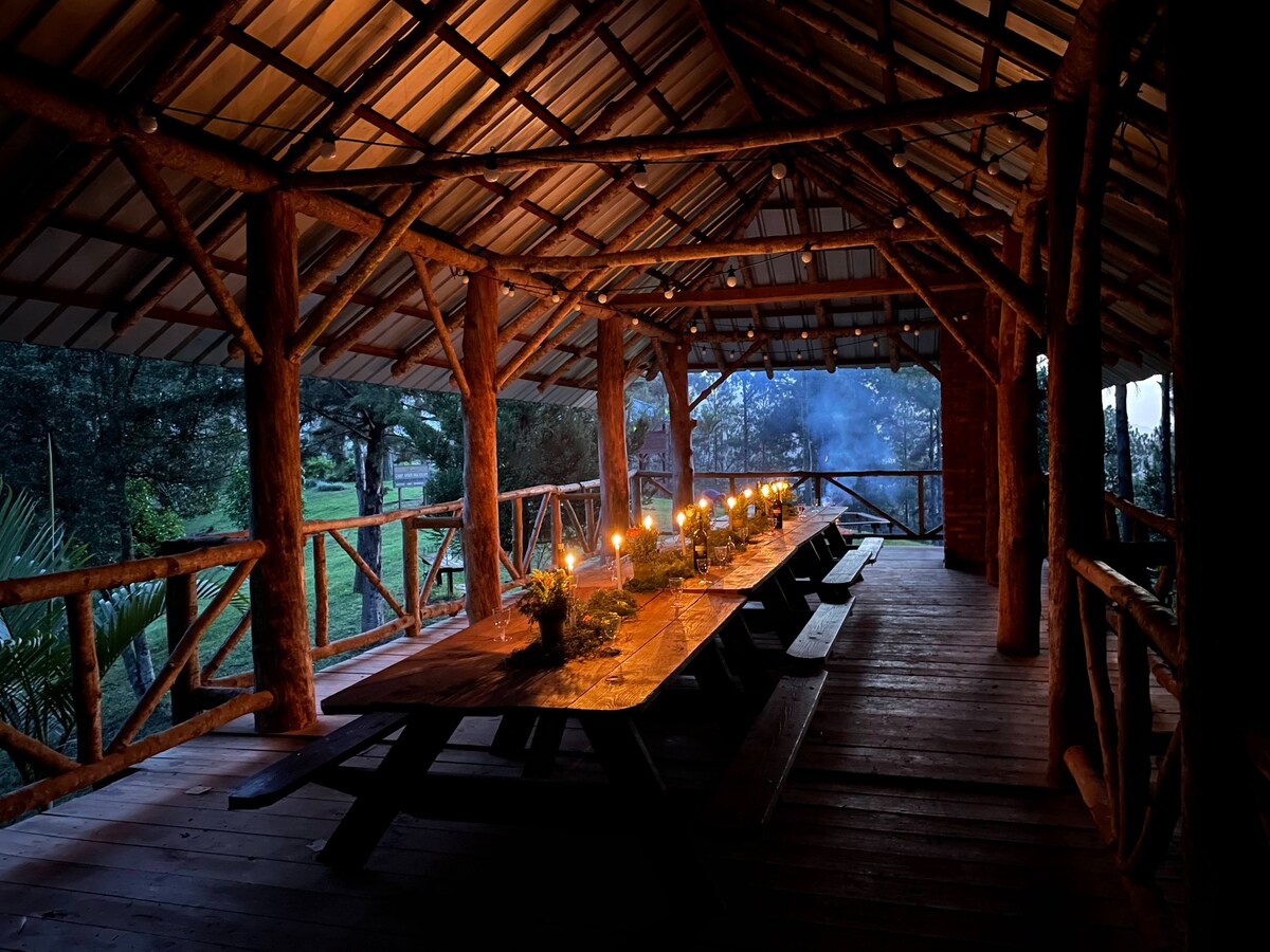 An open-air dining area features a long wooden table, softly illuminated by flickering candles. The structure is supported by rustic wooden beams, with surrounding views of the natural landscape creating a serene setting for gatherings.