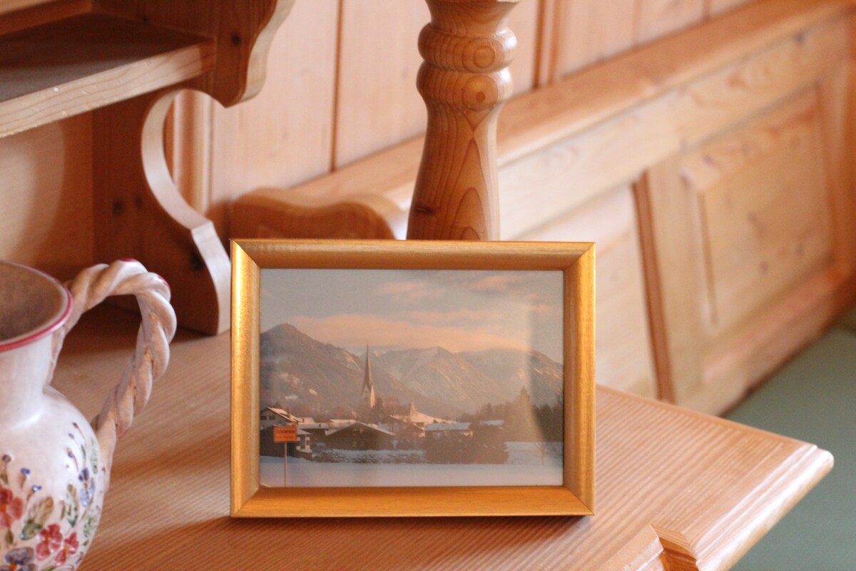 A gold-framed photograph is displayed on a wooden table. The image captures a winter scene featuring a quaint village and mountains in the background. Surrounding elements include a decorative ceramic pitcher and warm wooden surfaces, enhancing the room's rustic character.