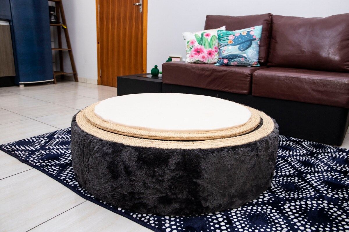 A round, soft ottoman in a dark grey fabric is placed on a patterned area rug. A light-colored circular cushion rests on top of the ottoman. In the background, a leather sofa with decorative pillows and a small shelving unit is visible.