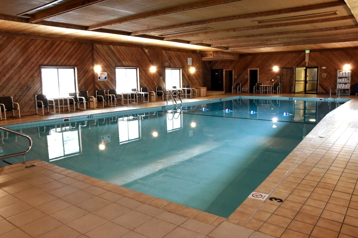 An indoor swimming pool is featured, with clear blue water spanning the area. Light reflects off the water's surface, while wooden paneling creates a warm atmosphere. Chairs are arranged around the poolside, and large windows provide natural light.