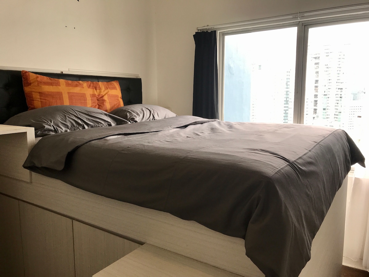 A cozy bedroom features a spacious bed with gray bedding and a decorative orange pillow. Natural light enters through large windows, offering a glimpse of the city skyline outside. Dark curtains are drawn to the side, complementing the modern room design.