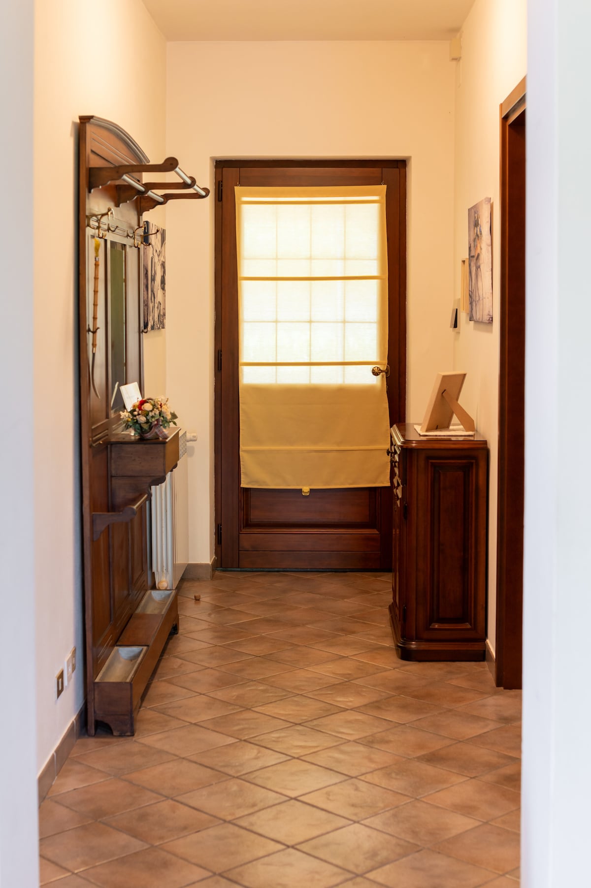 A narrow hallway is presented, featuring a wooden console table and a mirror on one side. The entrance door, partially covered with a light yellow curtain, allows diffused light to enter. Earth-toned floor tiles create a warm and inviting pathway to the interior.