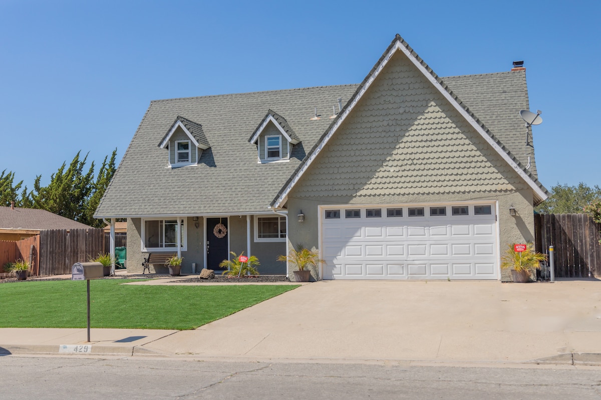 The exterior of a two-story house is showcased, featuring a pitched roof with decorative shingles. A well-manicured lawn is set in front, complemented by a driveway and a garage. Tall trees provide a backdrop, enhancing the peaceful setting.