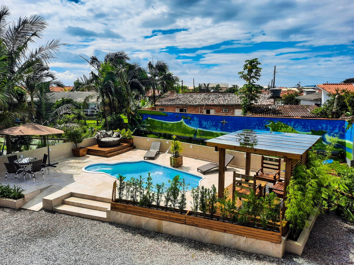 An outdoor area is highlighted by a small pool surrounded by sun loungers and shaded seating under a pergola. Lush greenery and a mural decorate the surroundings, adding a vibrant touch to the inviting space.