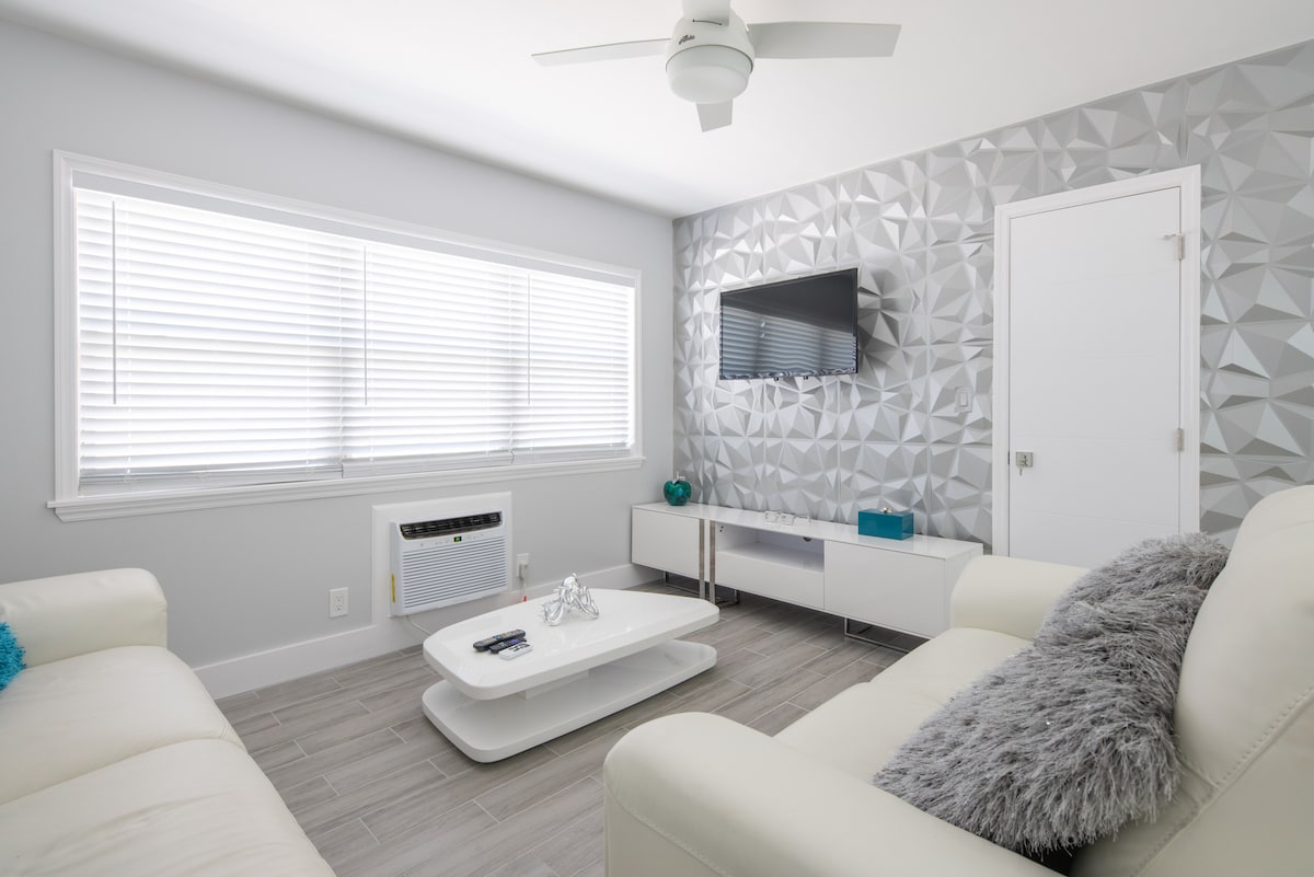 A modern living area features a white sectional sofa and a minimalist coffee table. A textured gray feature wall adds visual interest. Natural light filters through the blinds on two large windows, and a flat-screen TV is mounted for entertainment. A ceiling fan provides airflow.