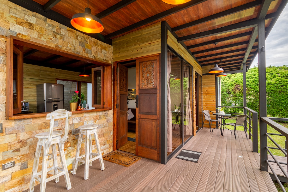 A covered porch features a natural wood and stone design, with tall glass doors leading to the interior. Two high stools are positioned at a bar area, while a small dining table and chairs await outside. Lush greenery is visible beyond the railing.
