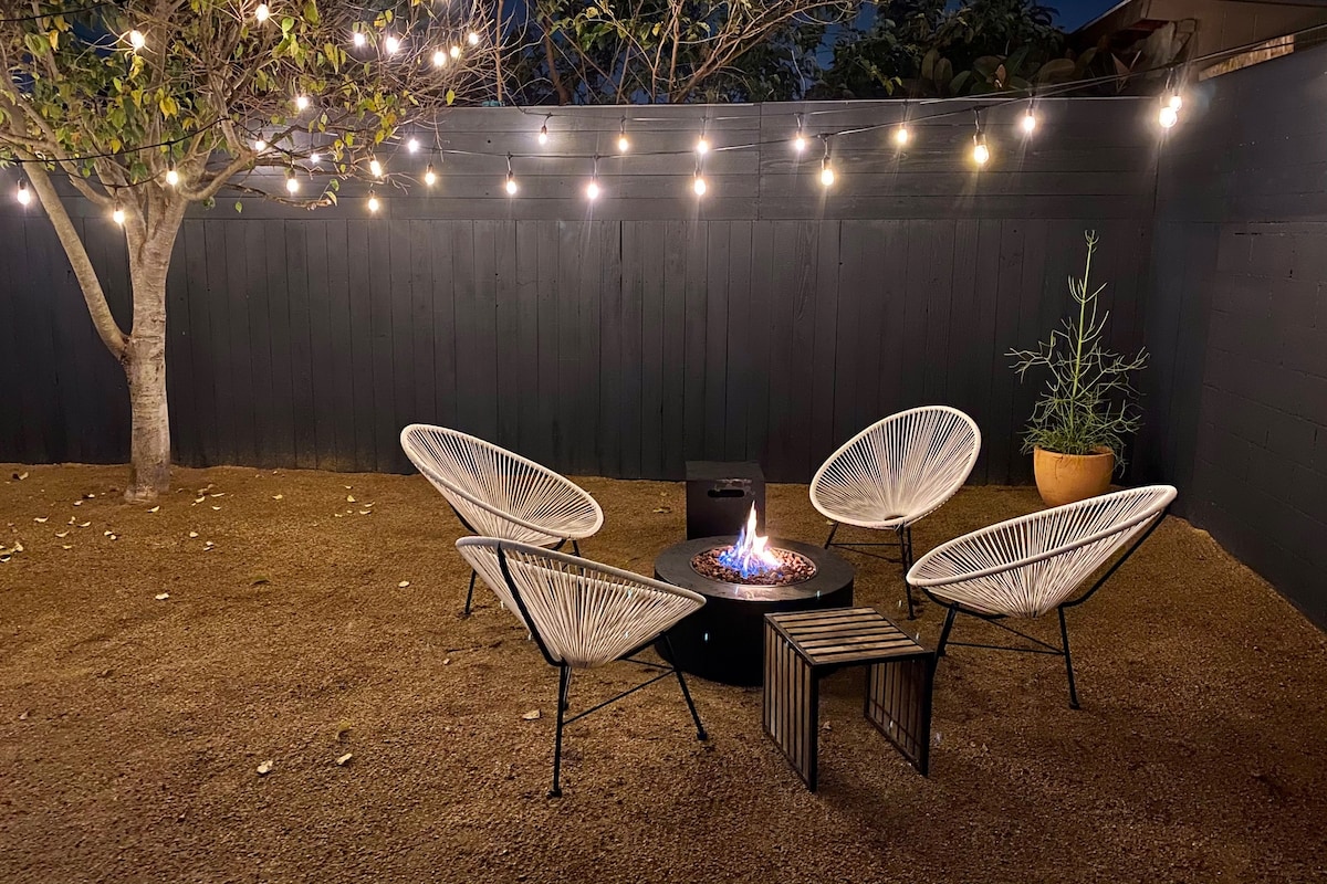 A cozy backyard setting is illuminated by twinkling string lights above. Four white lounge chairs are arranged around a circular fire pit, which features a gentle flame at the center. A small accent table sits beside one chair. A tree provides additional green accents in this relaxing space.