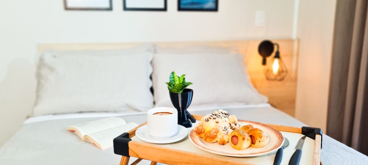 A breakfast tray is placed on the bed, featuring a cup of coffee and a plate of pastries. A small plant adds a touch of greenery. In the background, a neatly made bed with light bedding complements framed wall art.