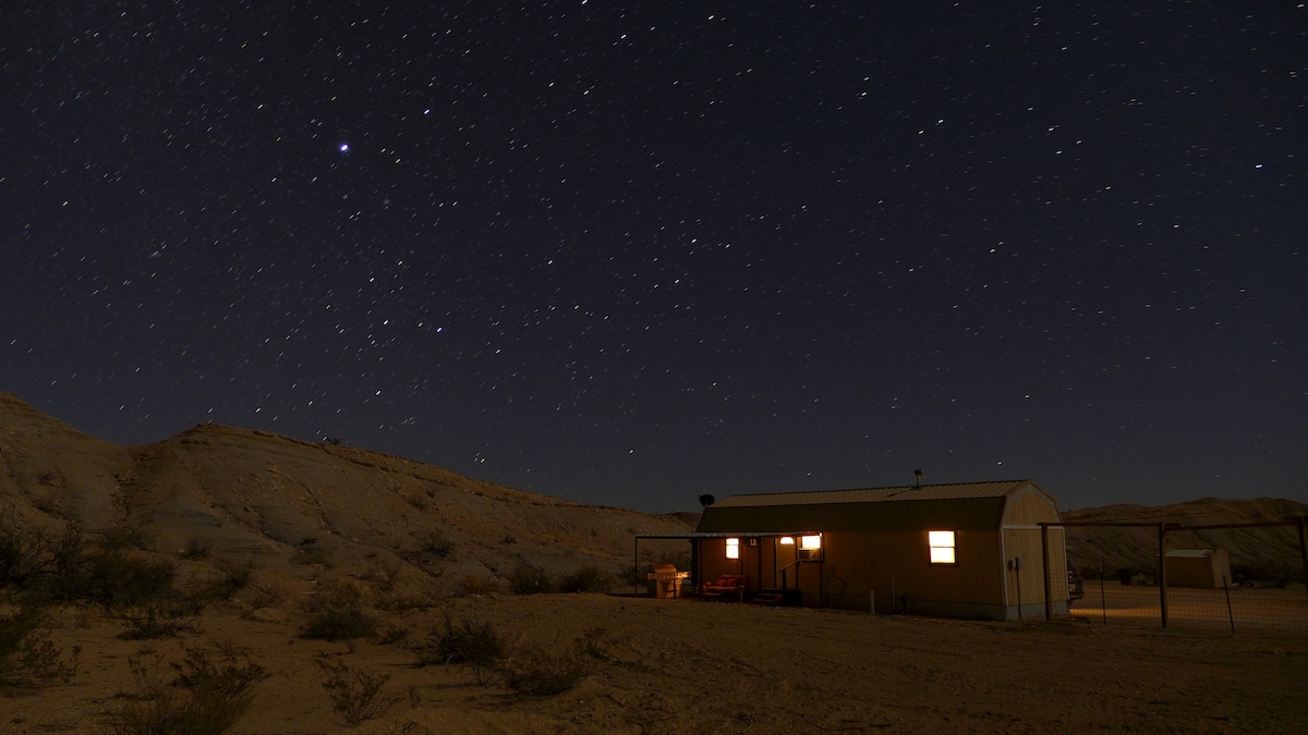A modest kabin is set against a backdrop of a clear night sky filled with stars. Soft light illuminates the windows, while the surrounding desert landscape features gentle hills and sparse vegetation.