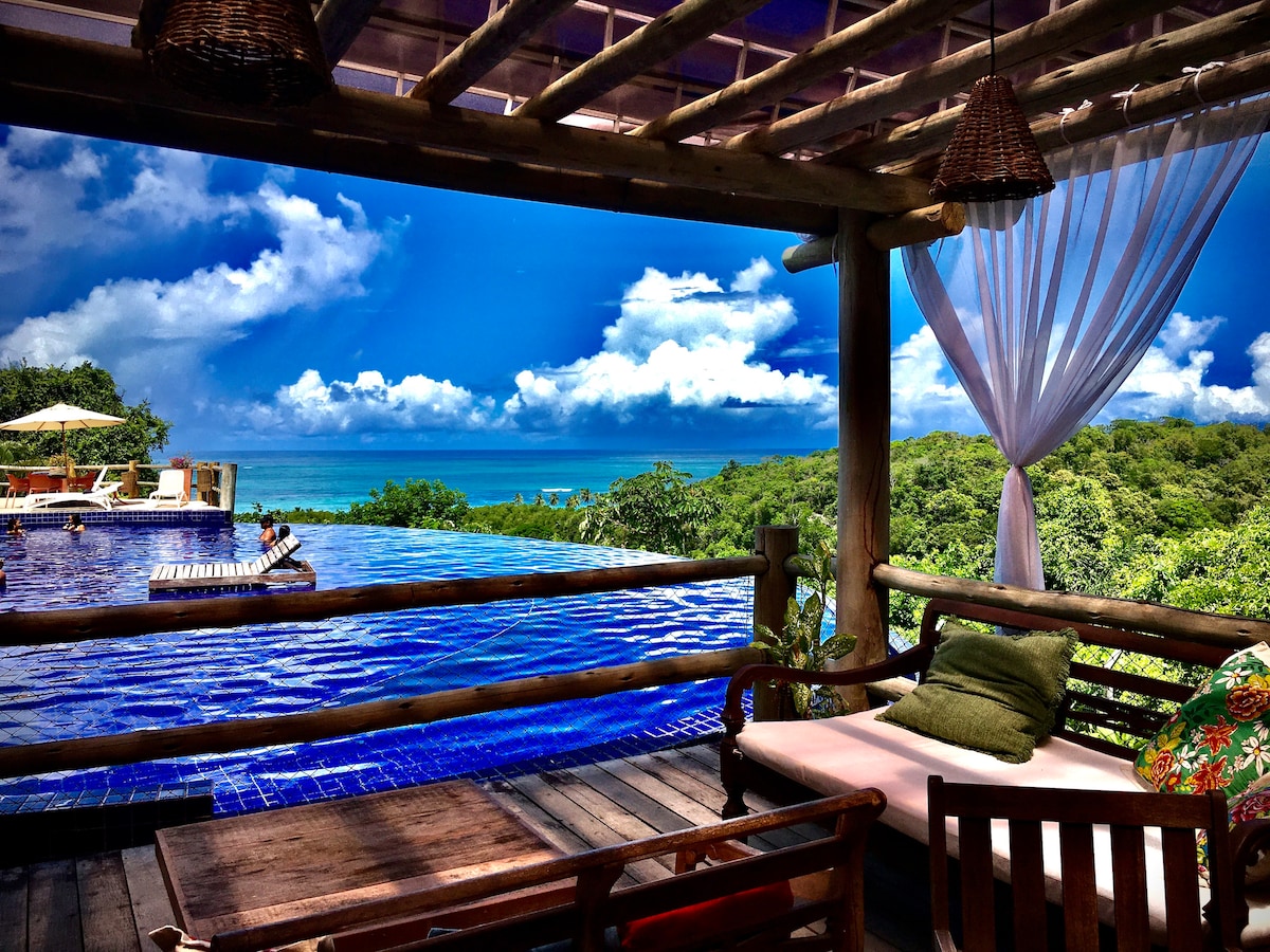 A spacious deck area is captured, featuring comfortable seating arrangements surrounded by lush greenery. An infinity pool provides a stunning view of the landscape and ocean beyond, with fluffy clouds visible in the blue sky. Sun loungers are positioned along the pool's edge, inviting relaxation.