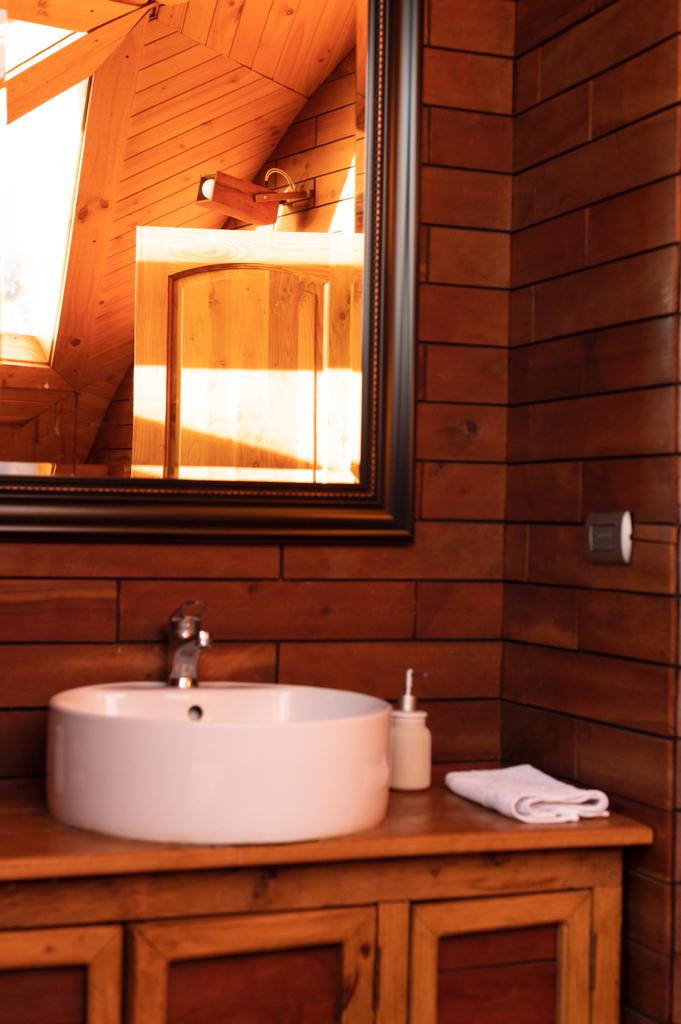A wooden bathroom features a round sink placed on a natural wooden countertop. A large mirror reflects the warm wood paneling and light coming from a nearby window. A neatly rolled towel and a soap dispenser are positioned beside the sink.
