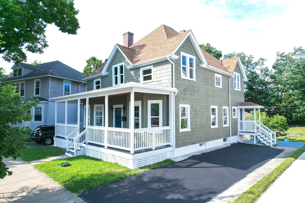 A spacious two-story home is shown, featuring a welcoming front porch with white railings and multiple oversized windows. The exterior is finished with light green siding, and a dark asphalt driveway provides ample parking space. A well-maintained lawn and surrounding trees add to the inviting ambiance.