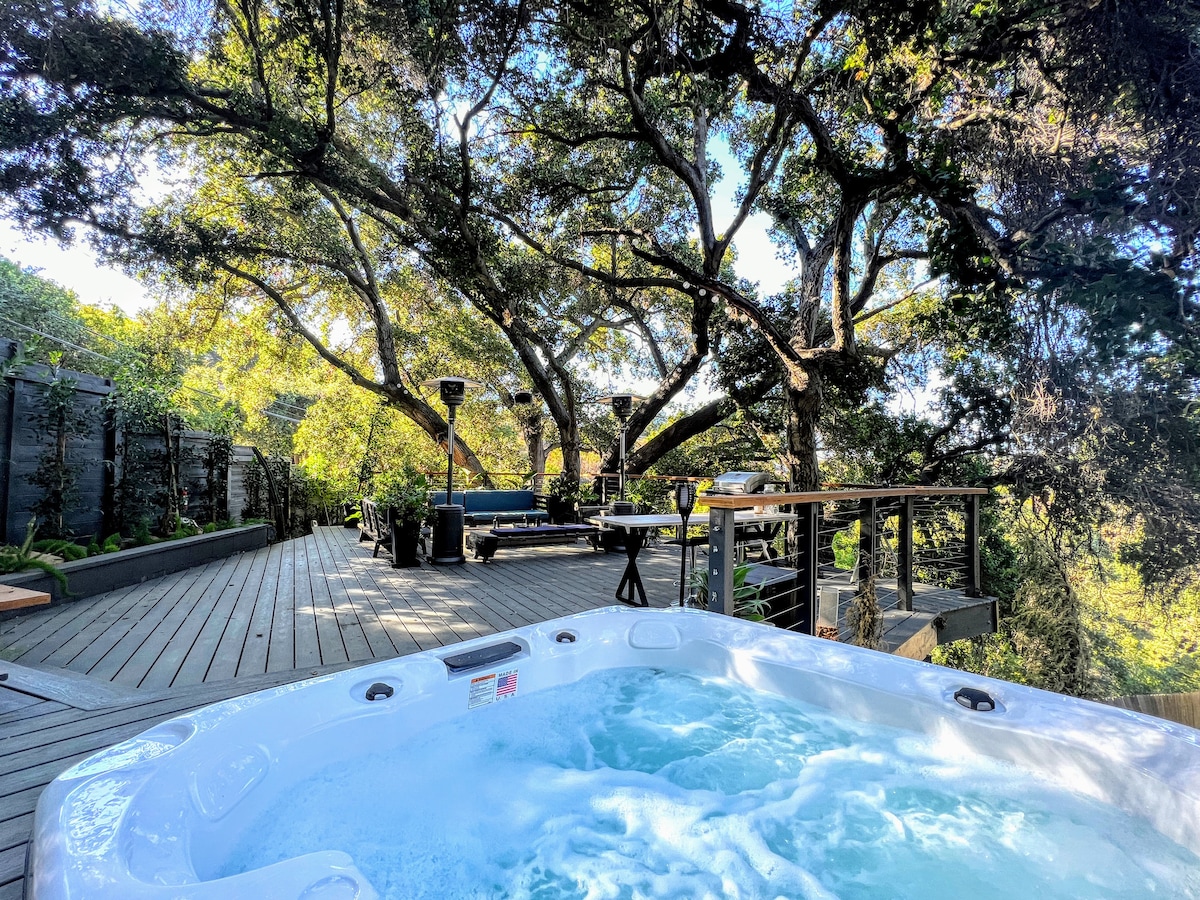 A hot tub provides a relaxing feature on the deck, surrounded by lush oak trees. In the background, an outdoor dining area is visible, with comfortable seating and tables set among greenery. The peaceful atmosphere is highlighted by dappled sunlight filtering through the leaves.