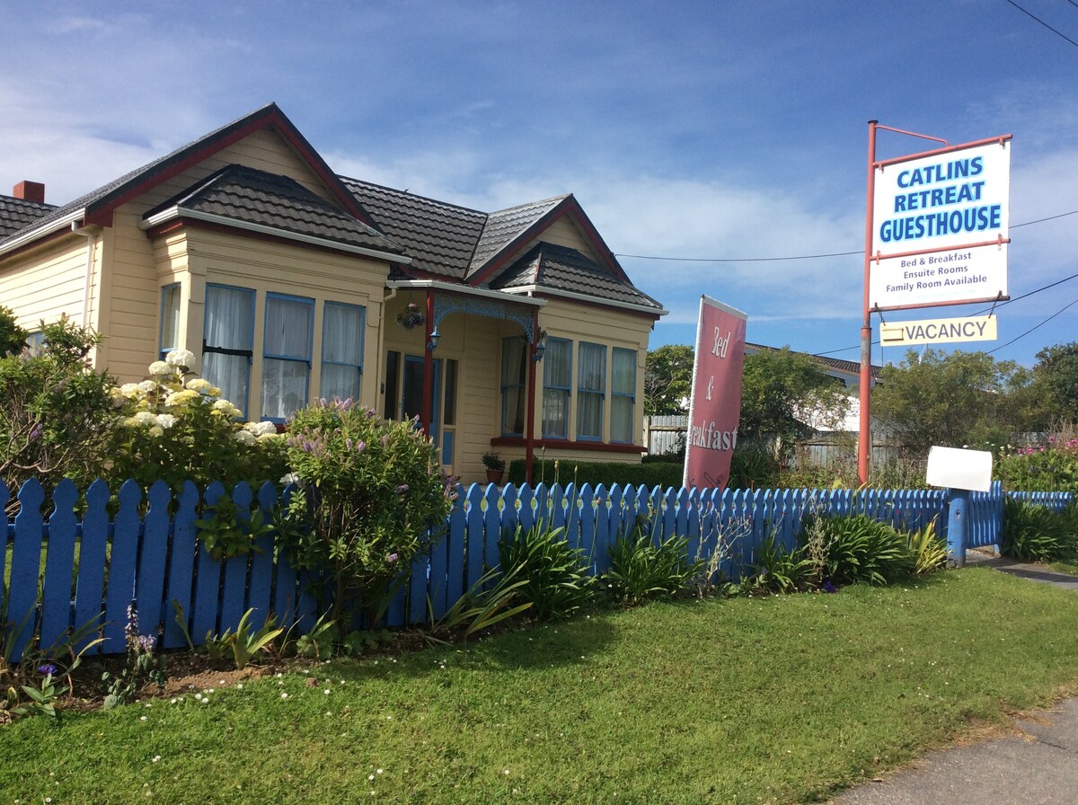 The historic Villa, Catlins Retreat, is marked by a welcoming sign and features a charming façade with blue trim. Lush greenery and flowering shrubs border the property, providing a vibrant appeal. A wooden fence encloses the yard, contributing to the inviting atmosphere of the guesthouse.
