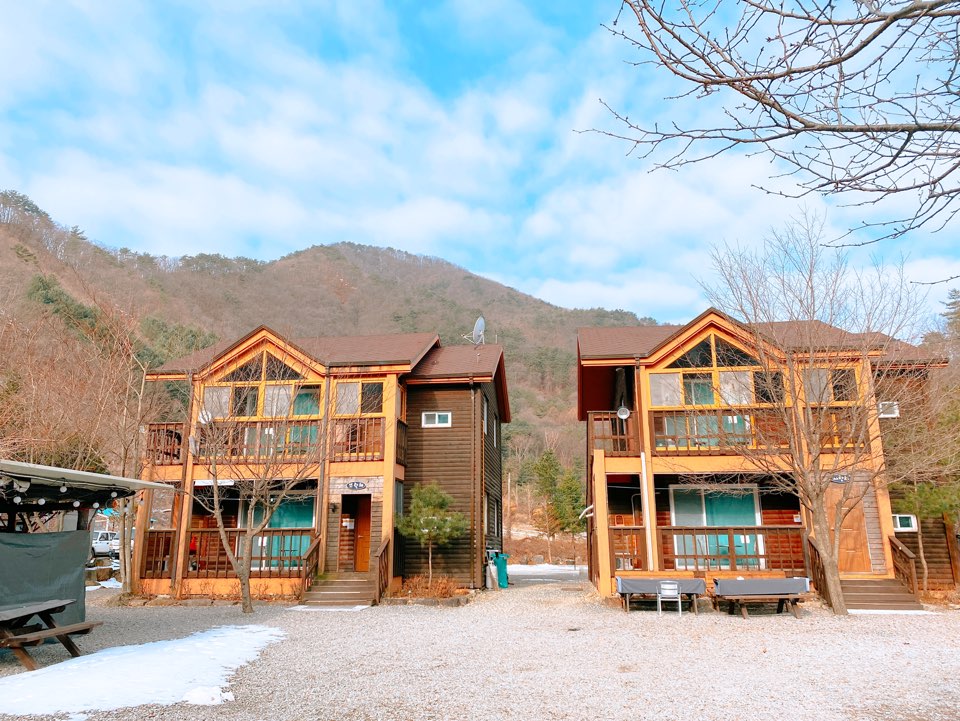 A charming two-story wooden building is viewed from the outdoor area, featuring a gravel pathway. Each unit has a balcony with wooden railings. Surrounding mountains are visible in the background, framed by bare trees, conveying a peaceful natural setting.