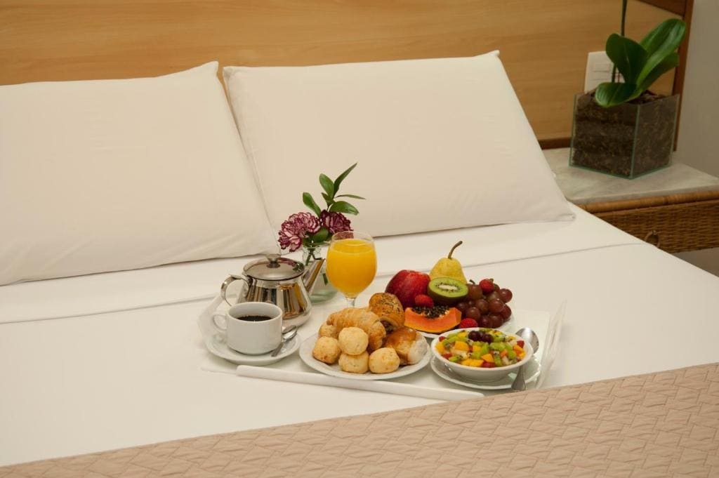 A tray is placed on a neatly made bed, showcasing a variety of breakfast items. A selection of pastries, fresh fruit, a glass of orange juice, and a cup of coffee are arranged alongside a small vase with a flower, adding a touch of decor.