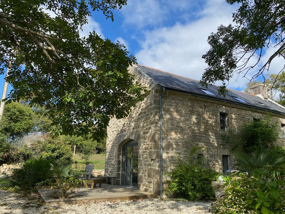 An ancient stone house is nestled beneath a canopy of trees, featuring a slate roof and large glass doors. A wooden deck with seating is visible in front, surrounded by greenery, while a clear blue sky can be seen above.
