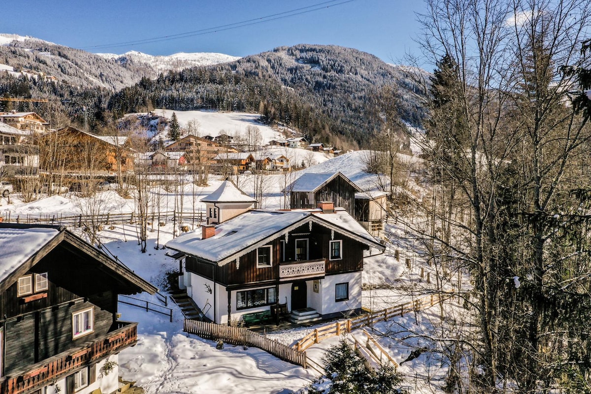 The exterior of the Alpine chalet is surrounded by a blanket of snow, with snow-covered roofs and a spacious yard. Towering mountains serve as a serene backdrop, while trees stand gracefully at the edges of the property, creating a picturesque winter landscape.