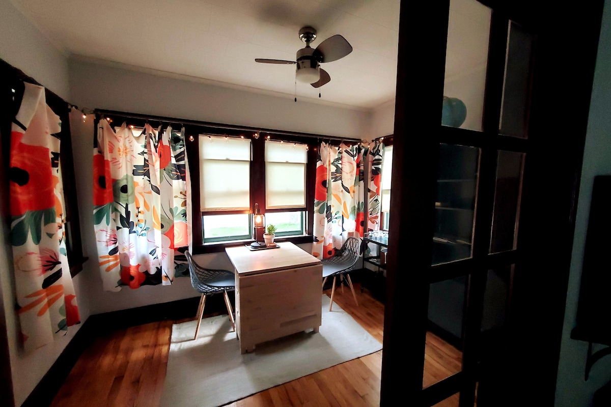 A bright and spacious room features large windows adorned with floral curtains, allowing natural light to filter through. A central table with two chairs sits on a light-colored rug, with a ceiling fan above providing a cool breeze.