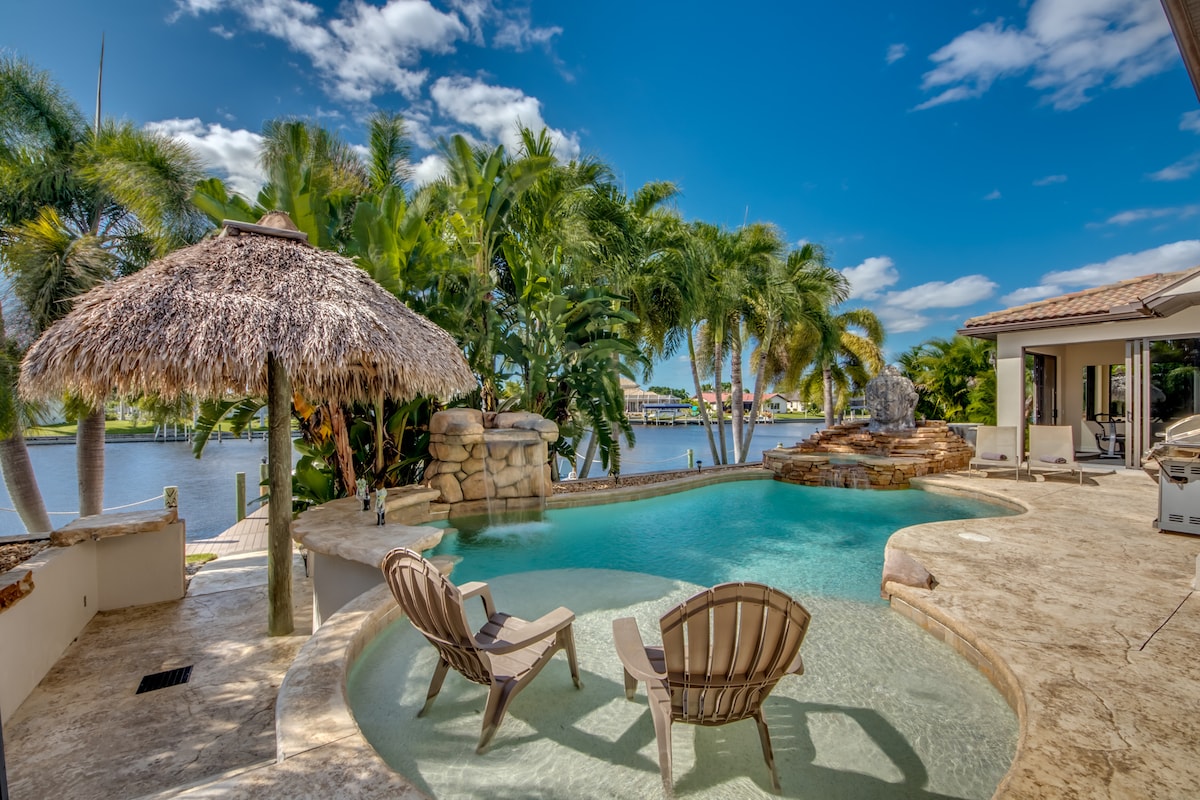 A serene outdoor space is featured, showcasing a tropical pool area with a thatched-roof Tiki hut. Two lounge chairs are positioned near the water, surrounded by lush palm trees. The crystal-clear pool reflects the blue sky and offers a scenic view of the canal.