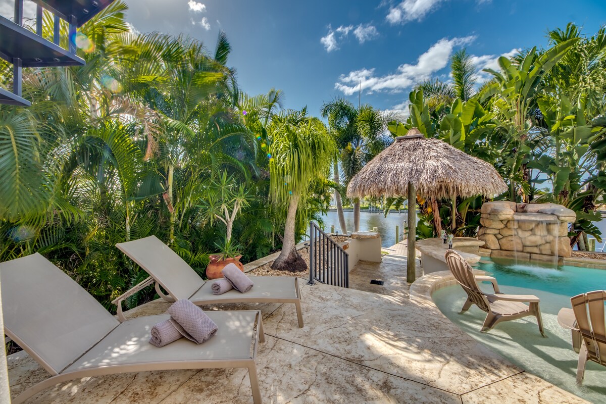 A serene outdoor area is surrounded by lush tropical plants, featuring a thatched-roof Tiki hut and a refreshing pool. Two lounge chairs are positioned near the water's edge, complemented by rolled towels. The scene captures a peaceful retreat with clear skies and vibrant greenery.