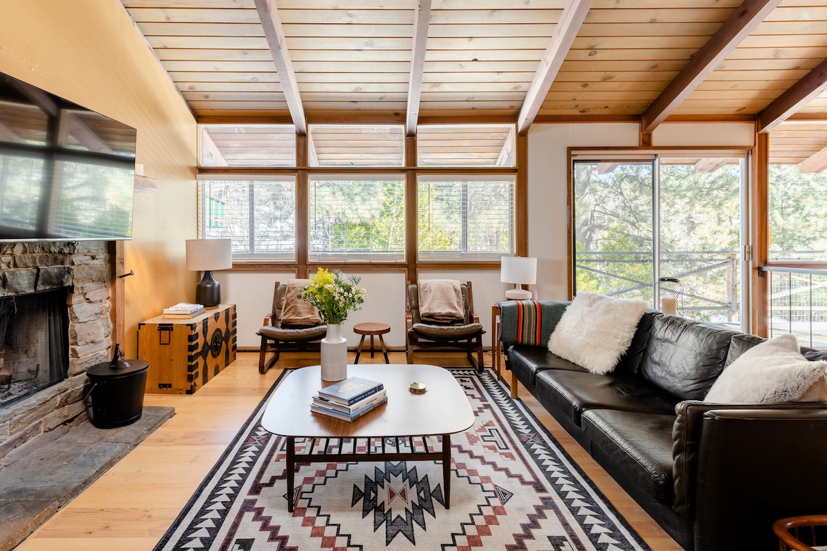 The living room features a wood burning fireplace framed by stone, with a comfortable black leather sofa and a round coffee table at its center. Large windows allow natural light to fill the space, showcasing views of surrounding trees. Two accent chairs offer additional seating.