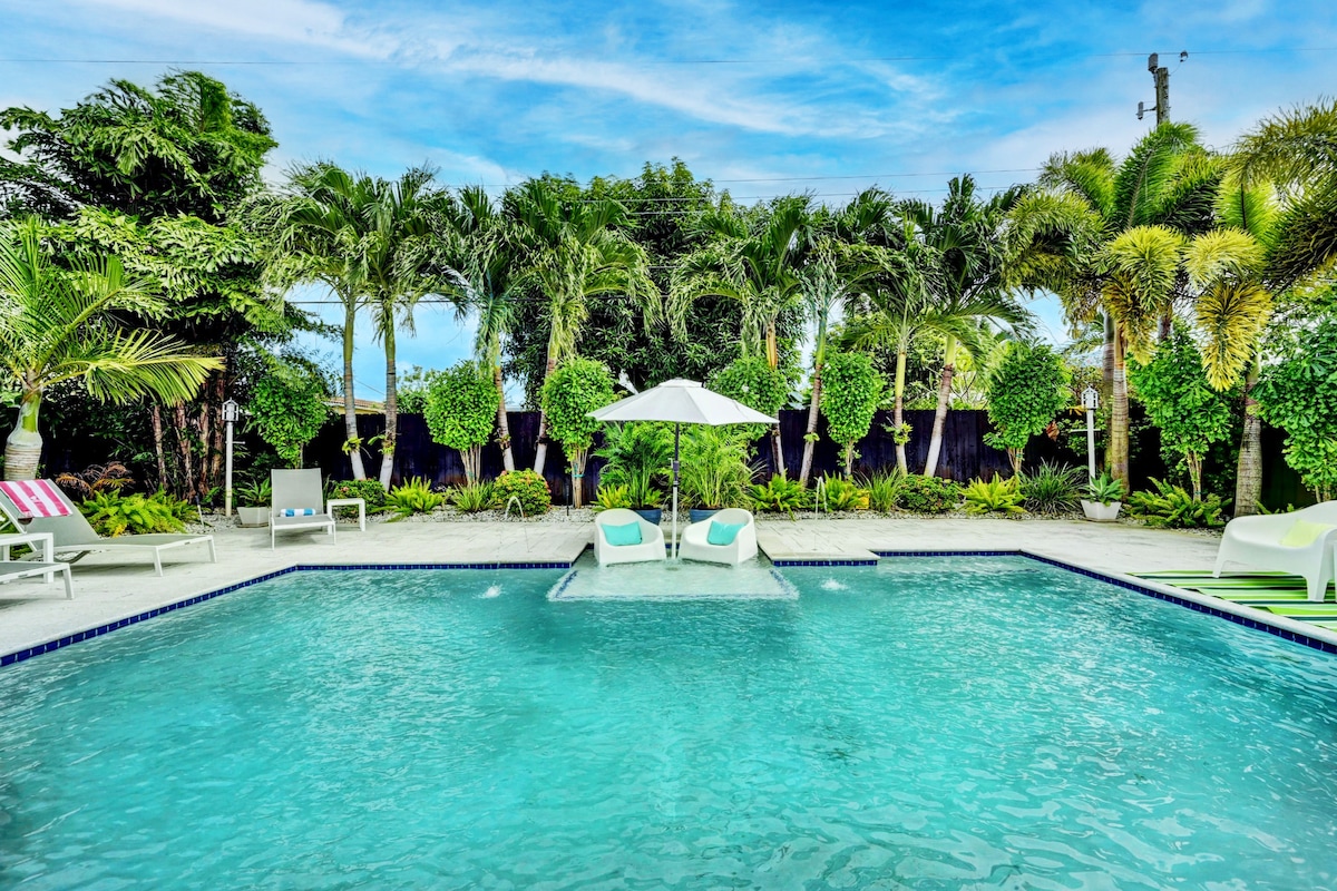 A spacious salt-water pool is surrounded by lush greenery and palm trees, creating a serene outdoor environment. A large umbrella provides shade over two lounge chairs positioned in the shallow end of the pool, inviting relaxation under the clear blue sky.