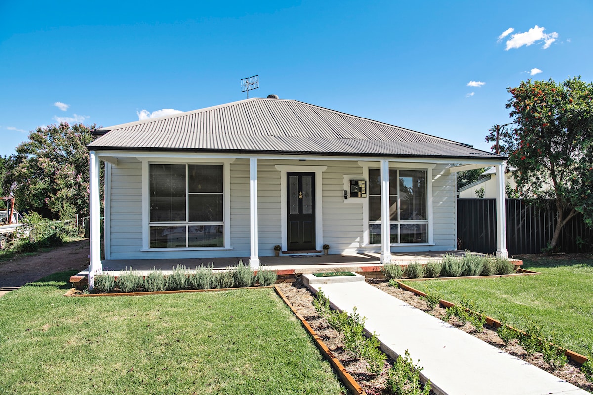 The exterior of the cottage showcases a light-colored façade with a classic design. A welcoming porch is framed by large windows on either side, allowing for ample natural light. The surrounding garden features neatly arranged greenery and a clear pathway leading to the entrance.
