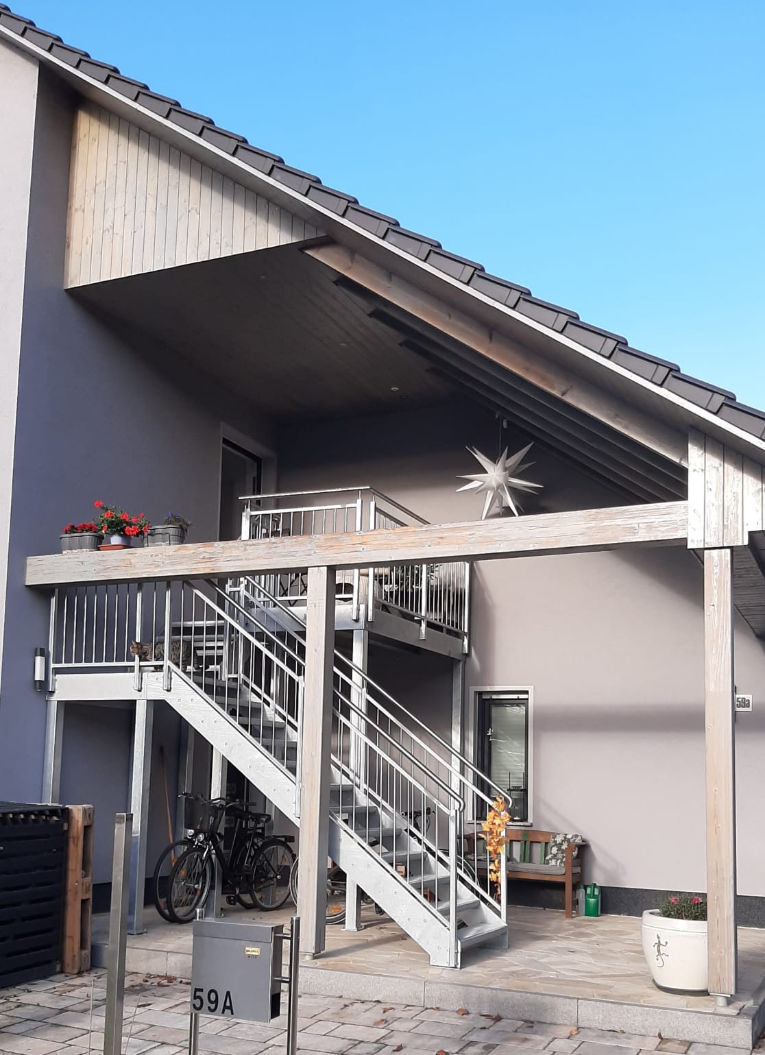 The entrance to the modern building is showcased with a structured exterior featuring a wooden balcony and decorative plants. A solid metal staircase leads to the upper level, while the exterior is complemented by a star-shaped ornament near the roofline.