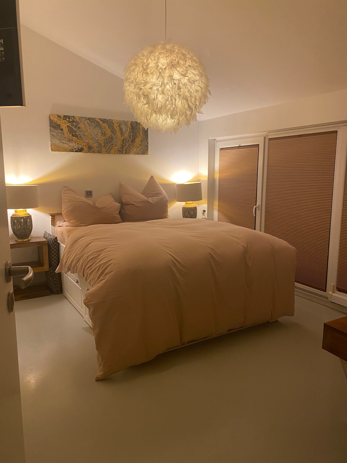 A cozy bedroom features a spacious bed with soft, neutral bedding and oversized pillows. Warm light is provided by two bedside lamps on matching tables, while a modern pendant light hangs above. Large glass doors lead to an outdoor area, complemented by soft window shades.
