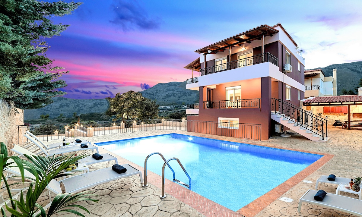 The villa is depicted with a multi-level design, surrounded by well-maintained outdoor spaces. A private pool is visible in the foreground, with sunbeds aligned nearby. Lush greenery and distant mountains create a serene backdrop, while soft colors painted across the sky indicate sunset.