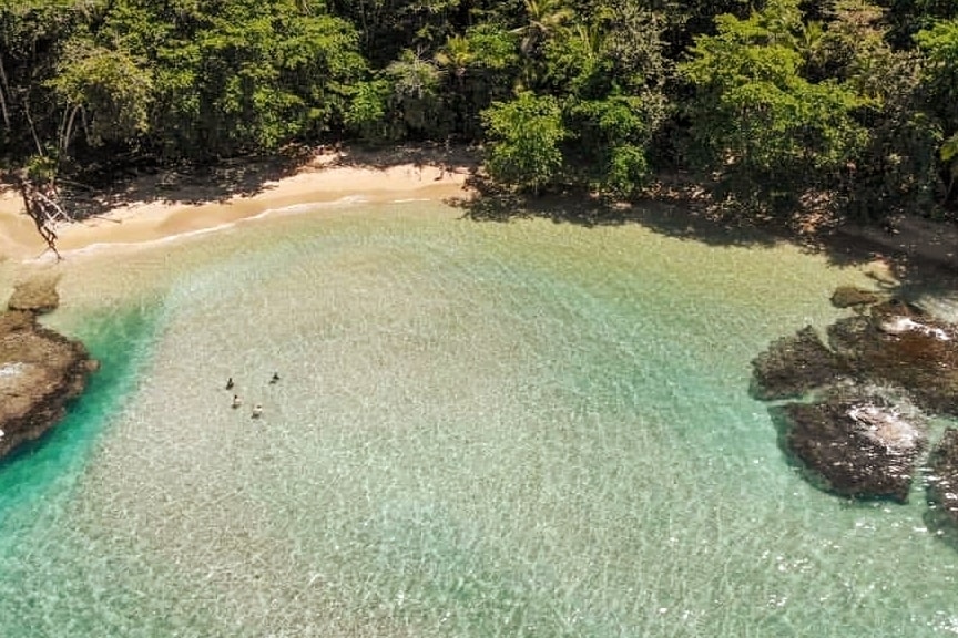 A secluded beach area features soft, golden sand bordered by lush green vegetation. Clear turquoise waters gently lap against the shore, with a few figures wading in the shallow water, enjoying the serene environment.