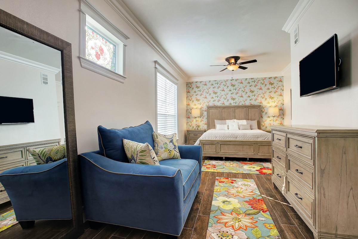 A spacious King bedroom features a cozy sitting area with a blue couch. Floral wallpaper adds a touch of color while a stylish area rug complements the wooden flooring. A large dresser and a flat-screen TV are positioned for convenience, creating a welcoming ambiance.