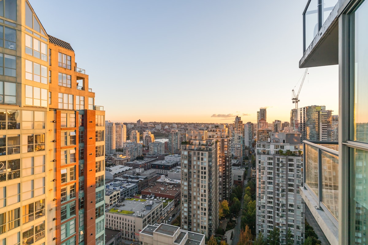 A stunning city skyline stretches across the horizon, with a blend of modern buildings reflecting the warm colors of sunset. The balcony offers unobstructed views of the vibrant urban landscape, highlighting the city's architectural variety and lively atmosphere.