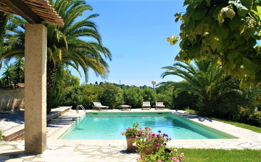 The swimming pool is surrounded by lush greenery and palm trees, offering a refreshing escape. Comfortable lounge chairs are positioned nearby for relaxing in the sun. Vibrant flowers in a pot add a colorful touch to the outdoor space, creating a serene atmosphere.